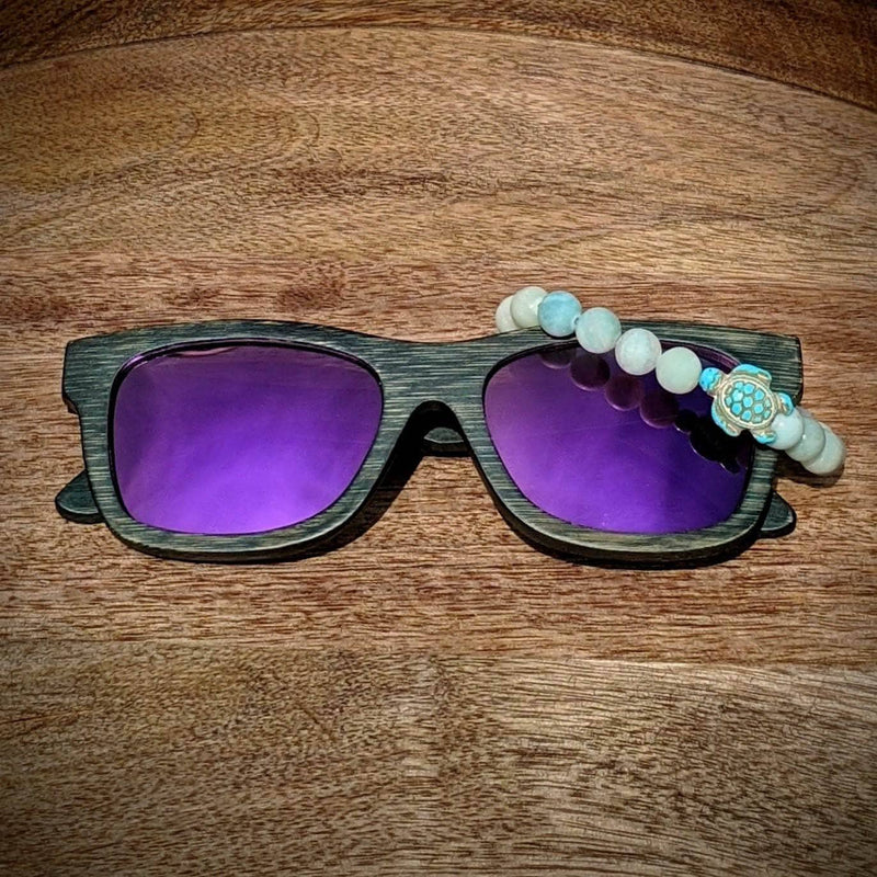 Purple lens dark bamboo sunglasses paired with a druzy agate turtle bracelet, displayed on a warm-toned wooden surface.
