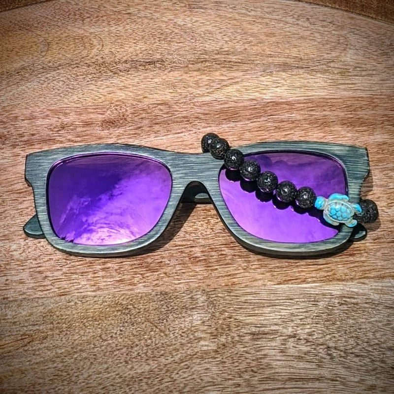 Purple lens dark bamboo sunglasses paired with a lave rock turtle bracelet, displayed on a rustic wood background.