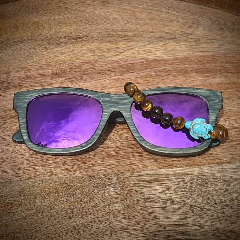 Purple lens dark bamboo sunglasses paired with a tiger’s eye turtle bracelet, displayed on a textured wooden background.
