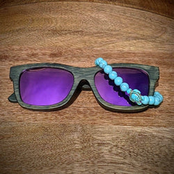 Dark bamboo sunglasses with purple mirror lenses paired with a turquoise gemstone turtle bracelet, displayed on a round wooden tray.
