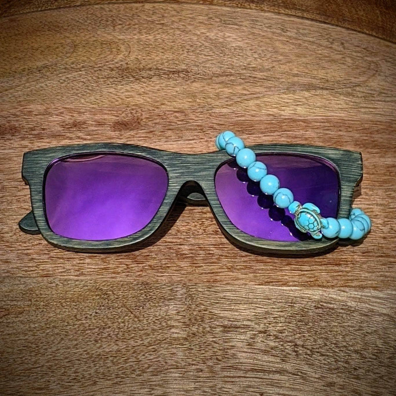 Dark bamboo sunglasses with purple mirror lenses paired with a turquoise gemstone turtle bracelet, displayed on a round wooden tray.
