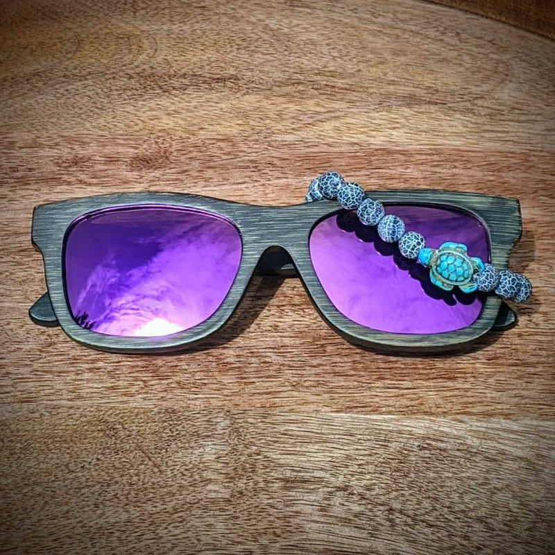 Purple lens dark bamboo sunglasses paired with a weathering agate turtle bracelet, displayed on a natural wooden surface.
