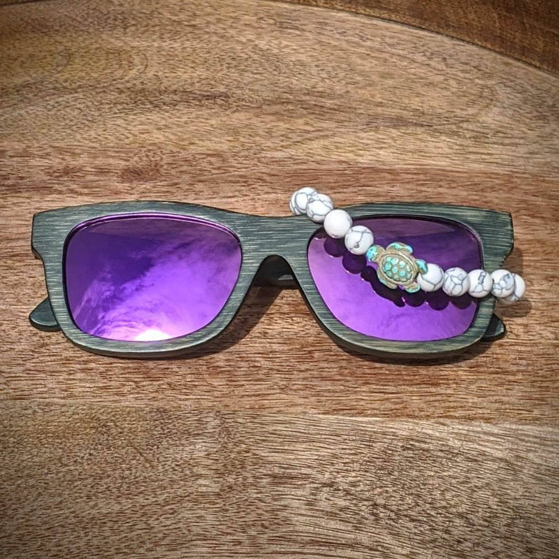 Purple lens bamboo sunglasses paired with a white turquoise gemstone turtle bracelet, displayed on a natural wooden surface.

