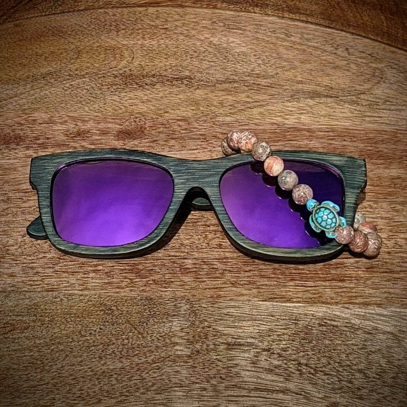 Black bamboo sunglasses with purple mirrored lenses and a rhodonite beaded bracelet featuring a turquoise turtle charm resting on wood.
