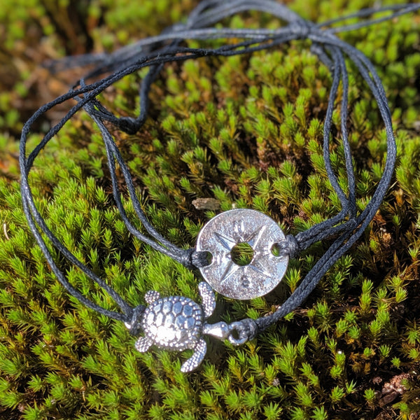 Silver compass and turtle charm bracelet on black adjustable cord, displayed on moss background.