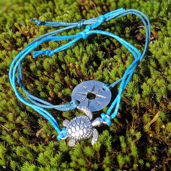 Blue cord bracelet with silver compass and turtle charms laid on green moss in natural light.