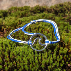 Adjustable blue and white cord bracelet with silver wave charm, displayed on moss in natural daylight.