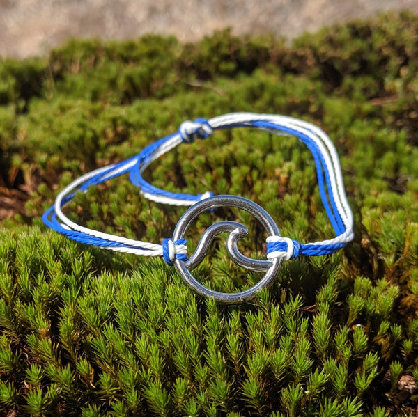 Adjustable blue and white cord bracelet with silver wave charm, displayed on moss in natural daylight.