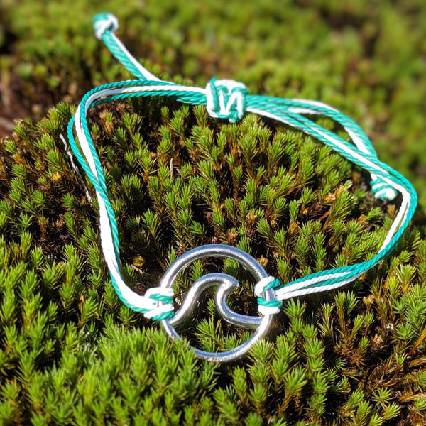 Silver wave charm bracelet on adjustable green and white cord, displayed on moss in natural light.
