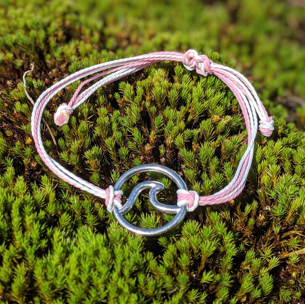  Silver wave charm bracelet on pink and white adjustable cord, laid on mossy surface in soft light.