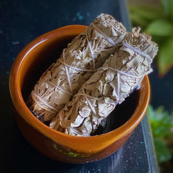 Two white sage smudge sticks in a terra cotta bowl on a natural wood surface.

