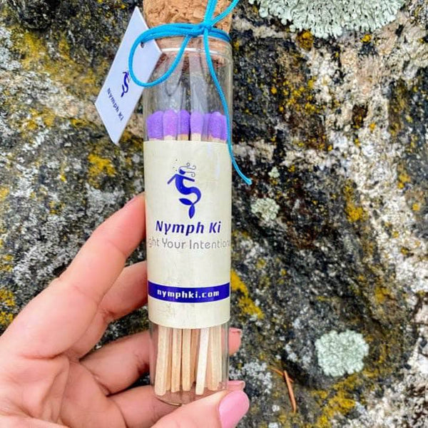 Glass ritual match jar with cork lid, purple-tipped matches, and Nymph Ki label, held by hand against a textured stone background.

