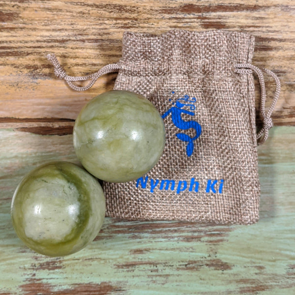 Two green jade Baoding balls placed on wood beside a Nymph KI drawstring pouch, designed for relaxation and meditation.